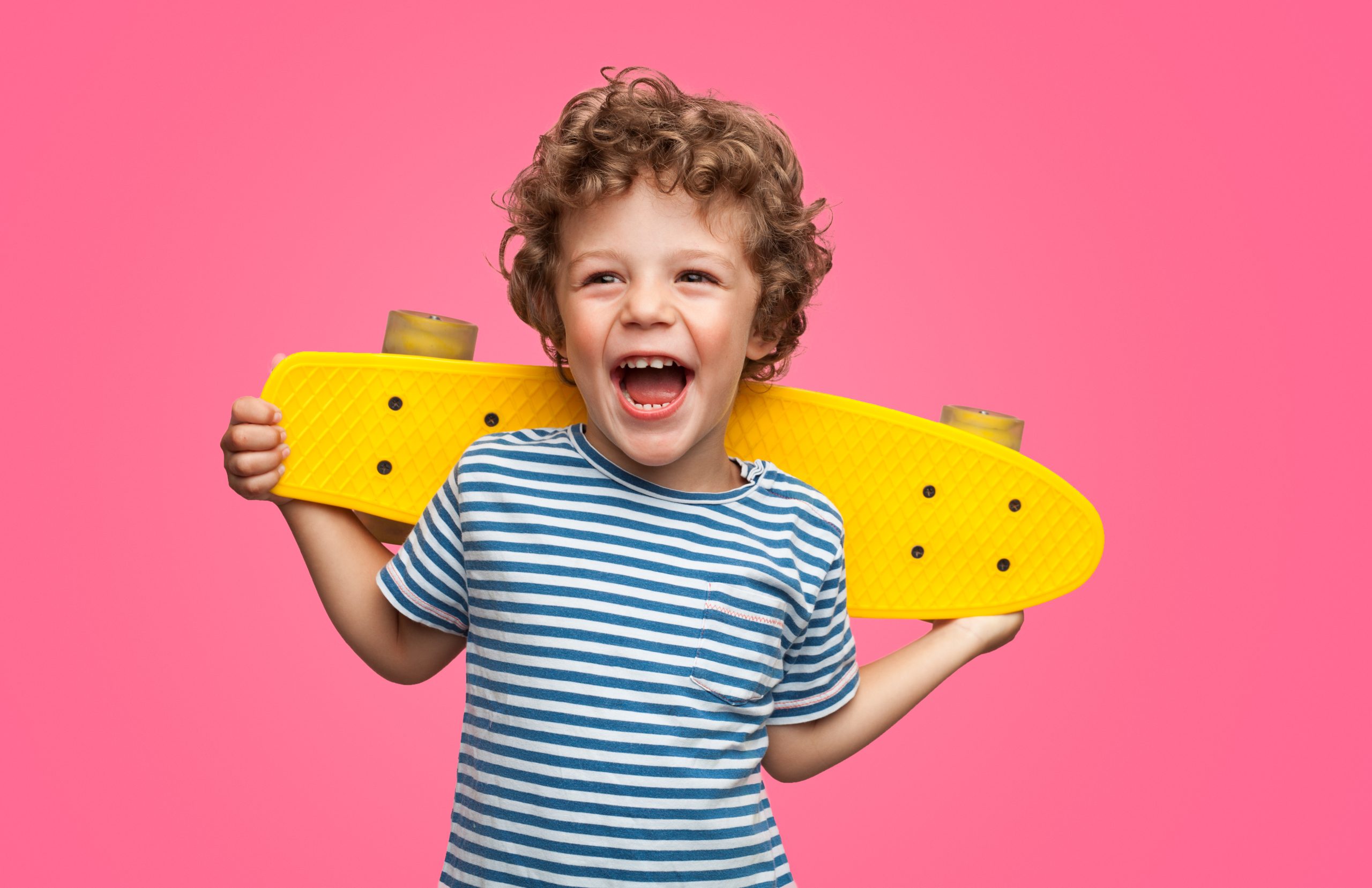 A very young curly haired child is holding a bright yellow skateboard across his shoulders. There is a pink background