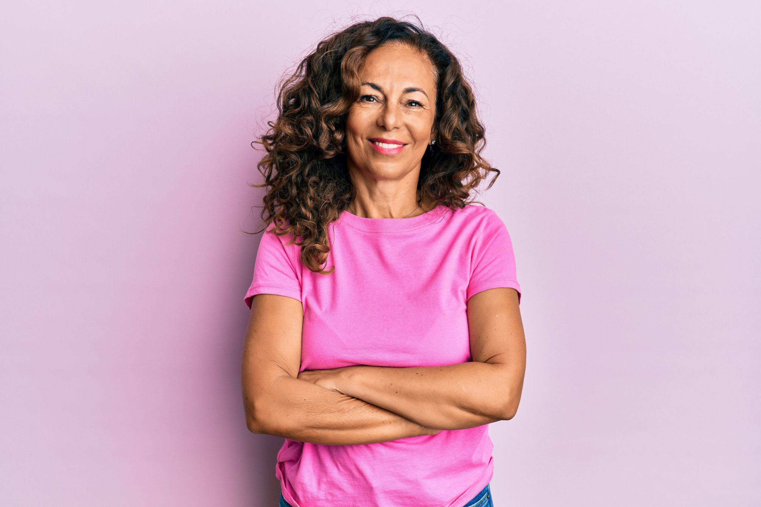A curly haired woman in a pink shirt is smiling with her arms crossed. She is standing in front of a light purple background