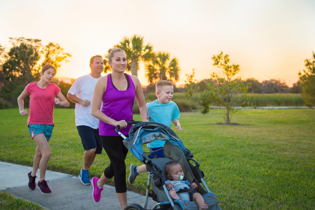 A young family is on an outdoor run by a park at sunset. The mother is pushing a stroller with an infant in it.