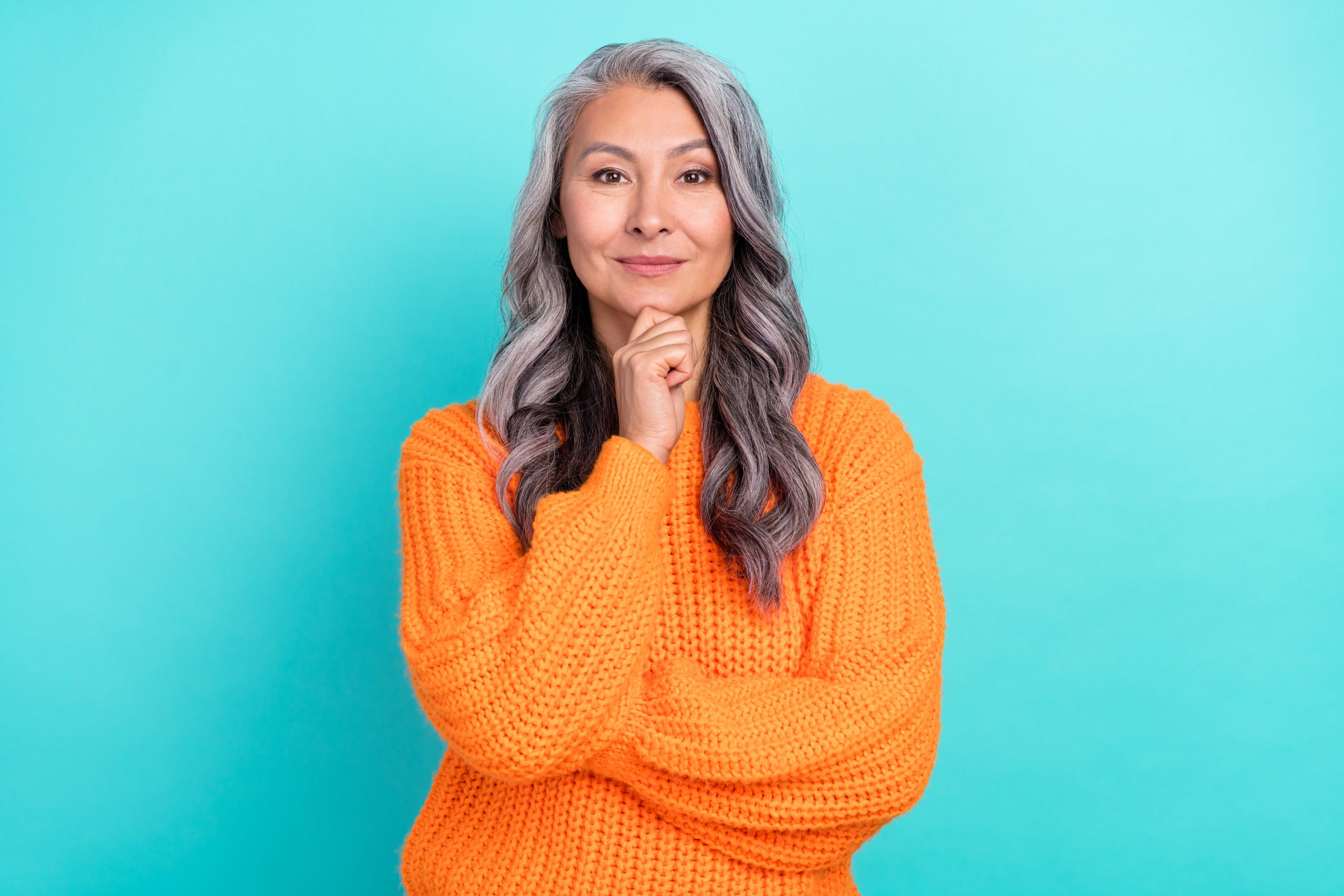 Portrait of a grey-haired woman with her hand on her chin standing in front of a teal turquoise background.