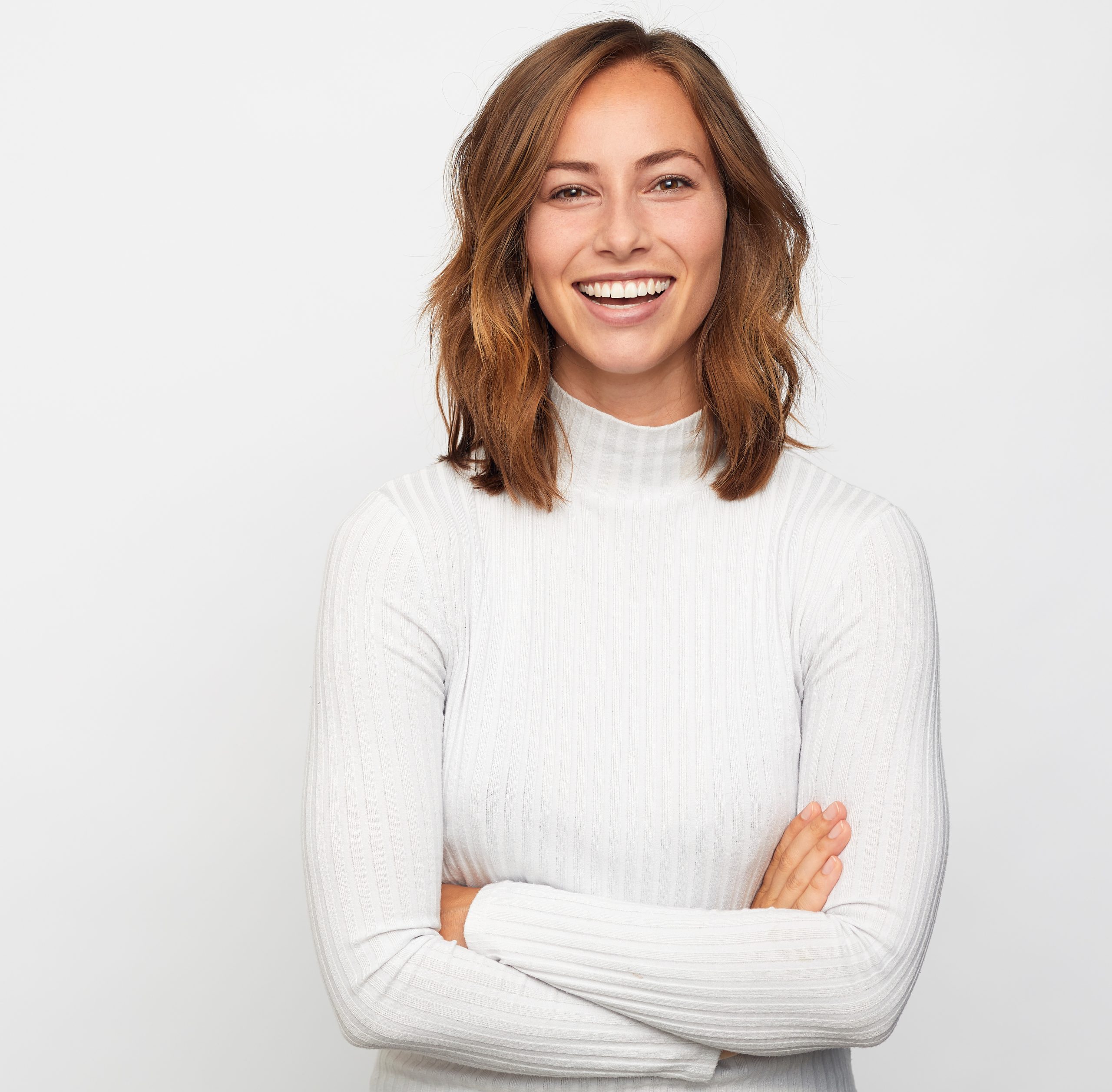 portrait of a smiling young woman in a white long sleeve shirt with her arms crossed in front of a light grey background
