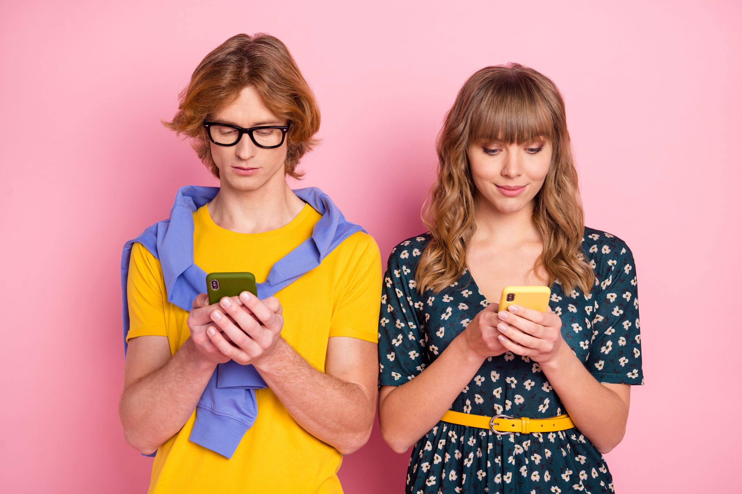 Two young people look down at their green and yellow smart phones while standing in front of a pink wall