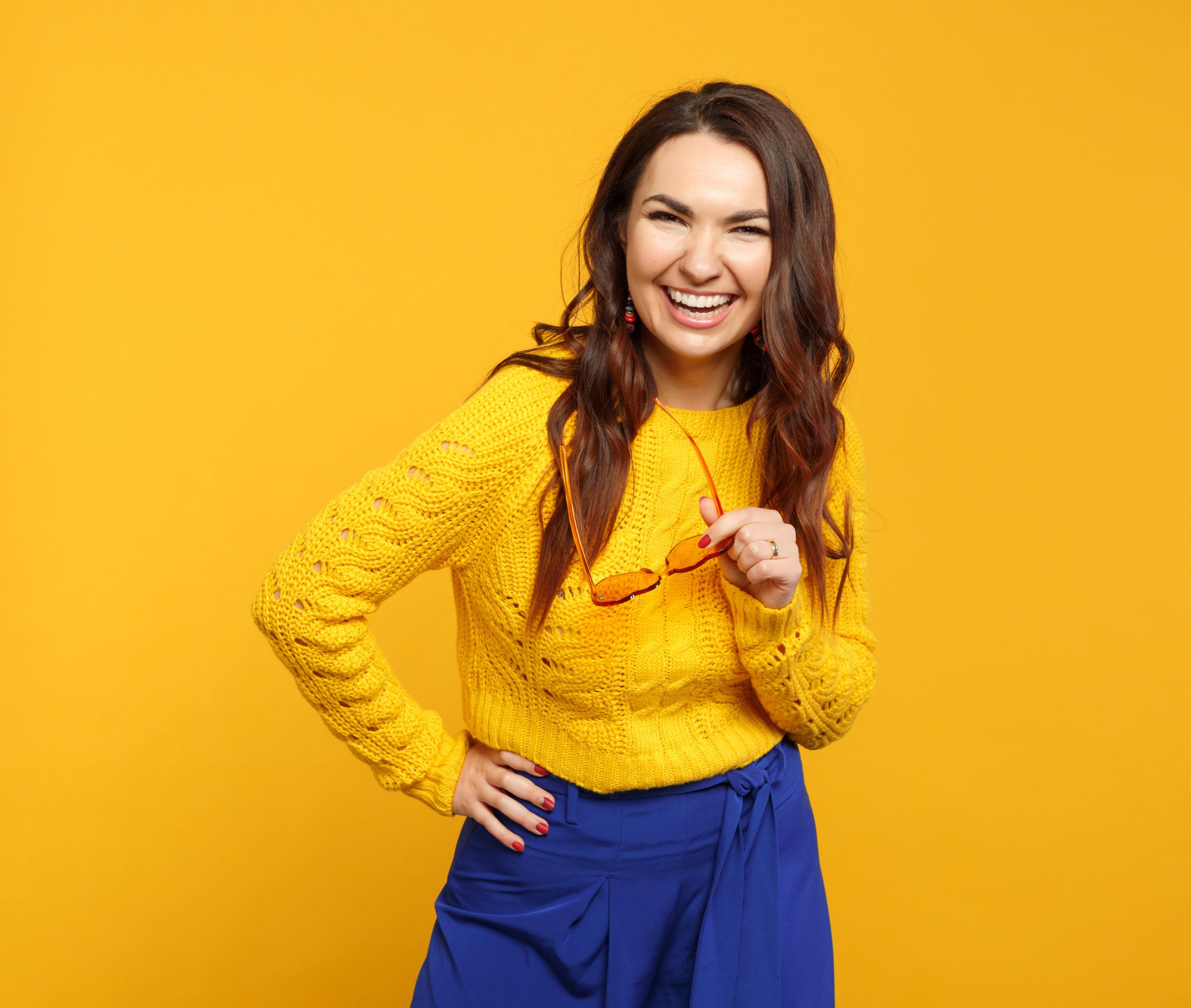 Portrait of laughing young woman in a yellow sweater and blue trousers holding glasses, standing, looking at the camera in front of a yellow-orange background