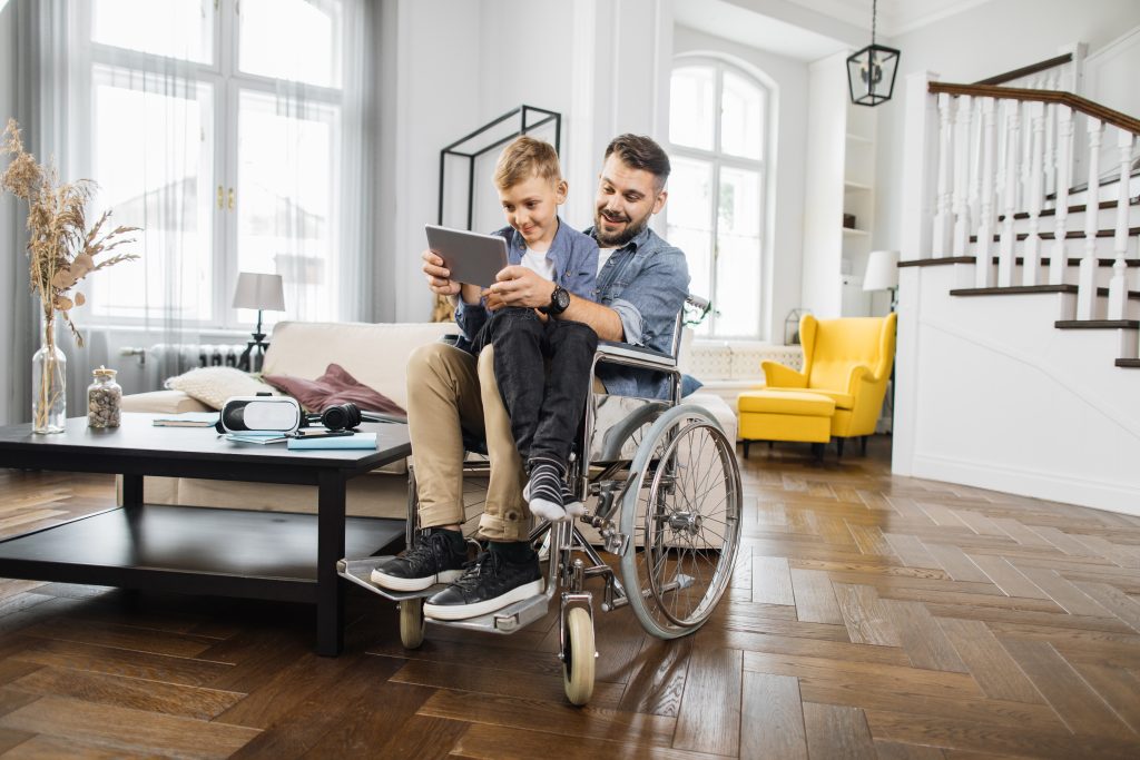 Man in wheelchair holding son and playing on tablet at home