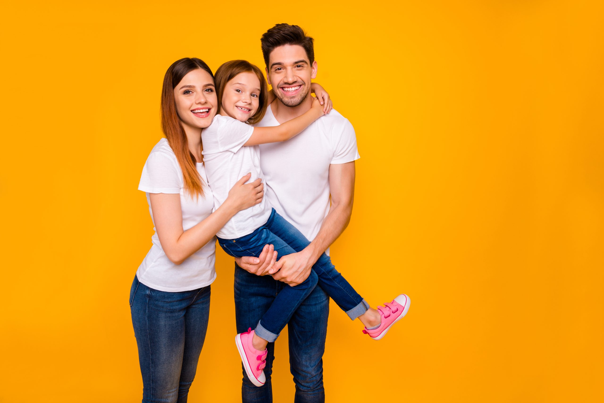 A family is posing for a photo. There is a mom, dad, and daughter all wearing white shirts and blue jeans. They are against a yellow background