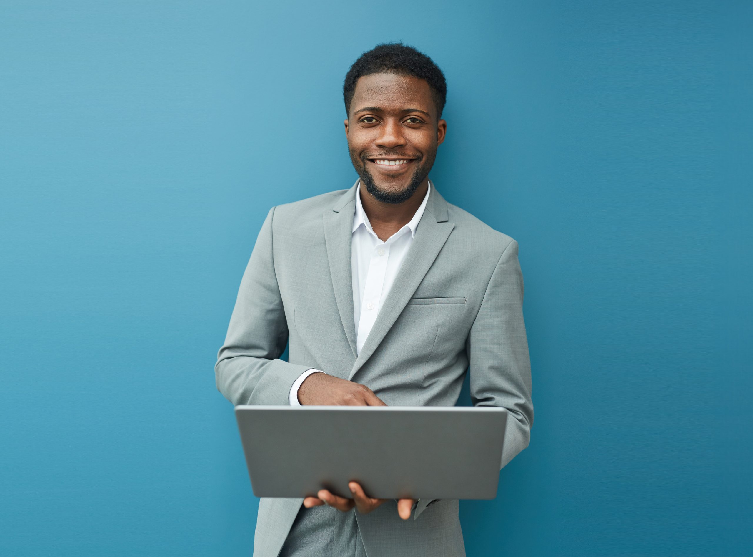 Portrait of smiling business man holding a laptop standing in front of a blue wall